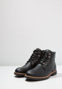 Botas de cuero negro hasta el tobillo con suela de goma gris, punta redonda y ojales de metal para los cordones. Incluye un logo grabado en el lateral.