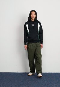 Musta huppari valkoisilla ja vihreillä yksityiskohdilla, joustavat hihansuut, yhdistettyinä löysiin oliivinvihreisiin housuihin ja valkoisiin tennareihin sinisellä matolla.