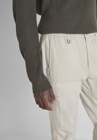 Tamnozeleni rebrasti pulover uparen s krem bojom cargo hlačama koje imaju bočne džepove i detalj s kukom blizu pasa.