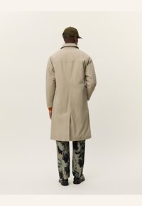 Palton bej cu o croială dreaptă și fante pe spate, purtat peste pantaloni cu model. Modelul poartă o șapcă închisă la culoare și mănuși cu accente portocalii.