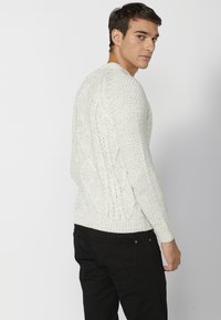 Weißer Strickpullover mit strukturiertem Rautenmuster, rundem Ausschnitt und gerippten Bündchen, kombiniert mit schwarzen Hosen.