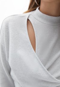 Ljusgrå stickad topp med hög hög krage, utskärningar vid axlarna och en omslag-design med en slät textur.