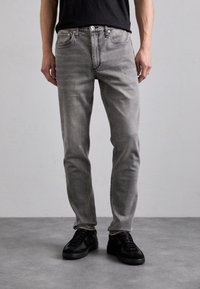 Jeans in denim grigio con vestibilità slim, design a cinque tasche, texture sbiadita e chiusura con bottone metallico, abbinati a sneakers nere.