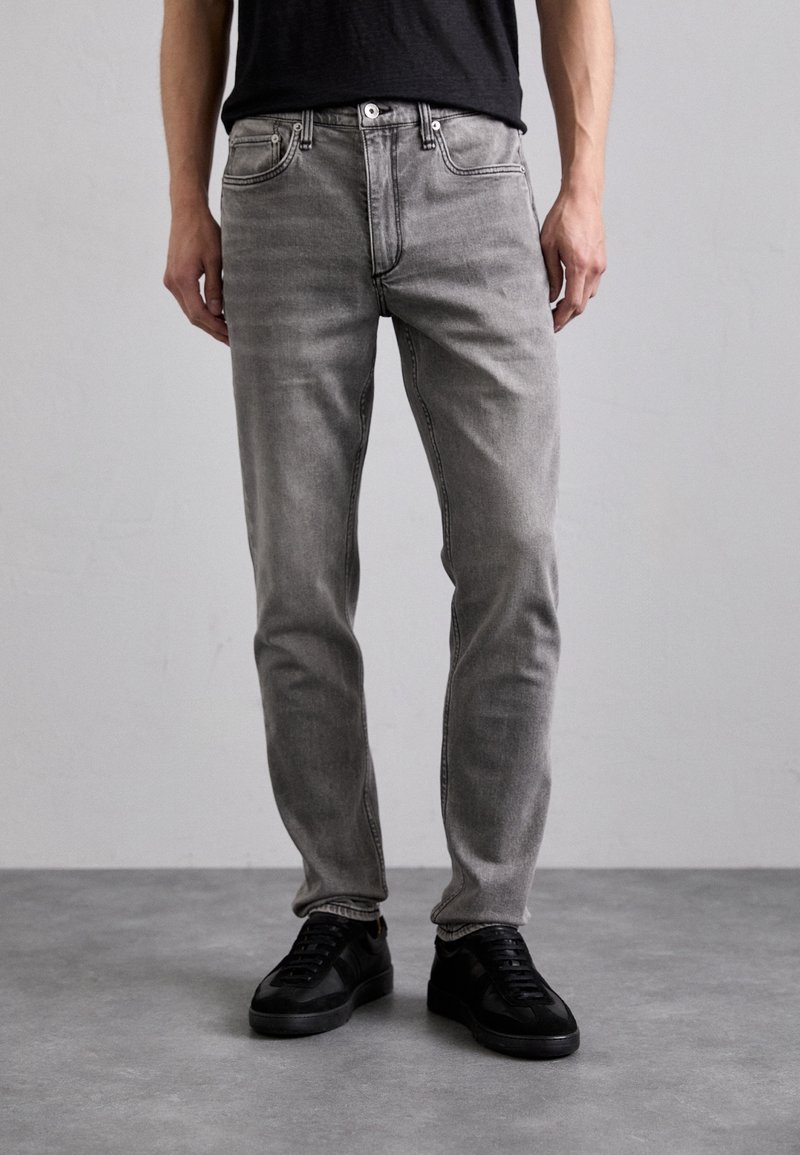 Jeans in denim grigio con vestibilità slim, design a cinque tasche, texture sbiadita e chiusura con bottone metallico, abbinati a sneakers nere.