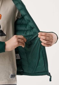 Mains tenant ouvertes la doublure intérieure d'une veste matelassée vert foncé, révélant la poche intérieure et la texture du tissu.