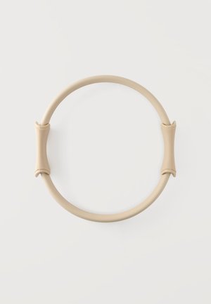 Cercle de pilates beige avec deux poignées courbées de chaque côté, posé sur un fond blanc uni.