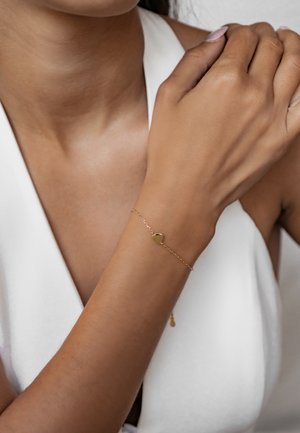Goldenes Armband mit einem herzförmigen Anhänger, zartes Kettendesign, getragen an einem braunhäutigen Handgelenk, vor einem weißen Hintergrund.