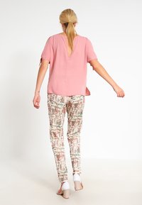 Světle růžový top s krátkým rukávem a výřezem na zádech, doplněný o kalhoty se vzorem geometrických tvarů v zemitých tónech a bílé sandály na bloku.