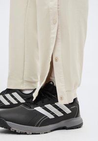 Μπεζ παντελόνι με ξεκουμπωτά κουμπιά στον αστράγαλο, φορεμένο με μαύρα και γκρι αθλητικά Adidas Traxion και λευκές κάλτσες.