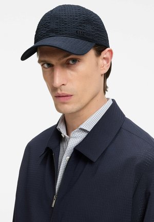 Mężczyzna o poważnym wyrazie twarzy nosi teksturowaną granatową czapkę i pasującą kurtkę na białej koszuli w wzory na jednolitym tle.