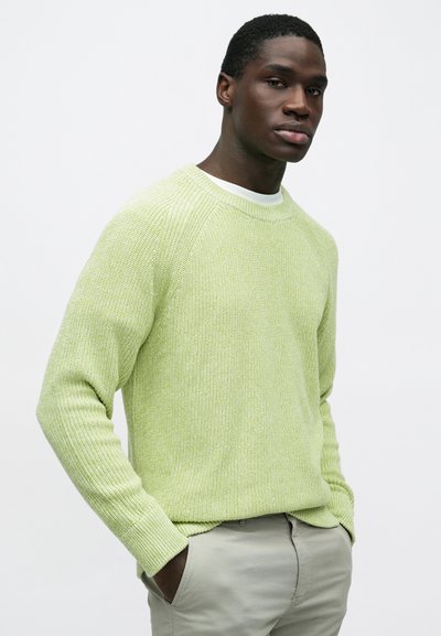 Mladý muž s rukami v kapse, oblečený v svetlozelenom pletenom svetri a svetlosivých nohaviciach na jednoduchom bielom pozadí.