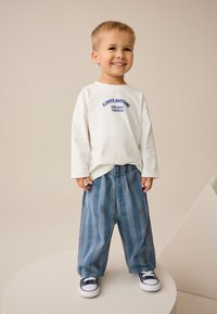 Vit långärmad skjorta med texten "ALWAYS AWESOME", kombinerad med blå randiga byxor. Barnet bär vita sneakers och står på en rund plattform.