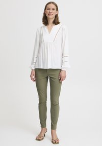 Weißes Blusenshirt mit V-Ausschnitt und puffärmel, mit Lochdetails. Kombiniert mit olivgrünen Slim-Fit-Hosen und grünen Absatzsandalen.