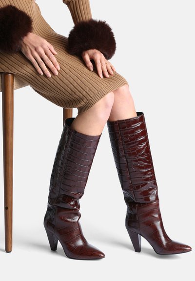 Femme assise sur un tabouret en bois portant une robe marron côtelée avec des poignets en fourrure foncée et des bottes à talons hauteur genou marron foncé à motif crocodile.