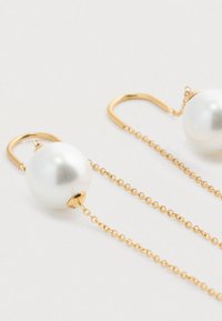 Finom arany lánc fülbevalók nagy fehér gyöngyökkel és arany kampós zárakkal, egy világos felületen pihenve.