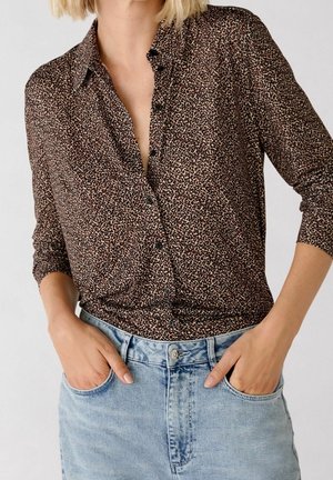 Femme portant un chemisier marron à manches longues à imprimé léopard, rentré dans un jean taille haute bleu clair, les mains dans les poches.