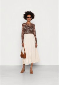 Mulher vestindo uma blusa de manga longa com padrão de leopardo, saia midi plisada em creme, óculos de sol, bolsa castanha e sandálias de salto, em pé contra um fundo branco.