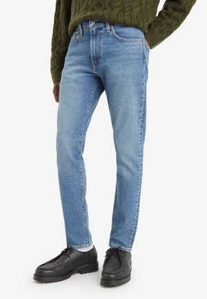 Man draagt lichtblauwe jeans met rechte pijpen, witte sokken, zwarte veterlaarzen en een olijfgroene kabelgebreide trui.