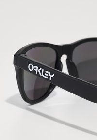 Ochelari de soare negri cu lentile întunecate, având un finisaj mat. Bratul prezintă un logo alb "Oakley". Design cu un cadru unghiular.