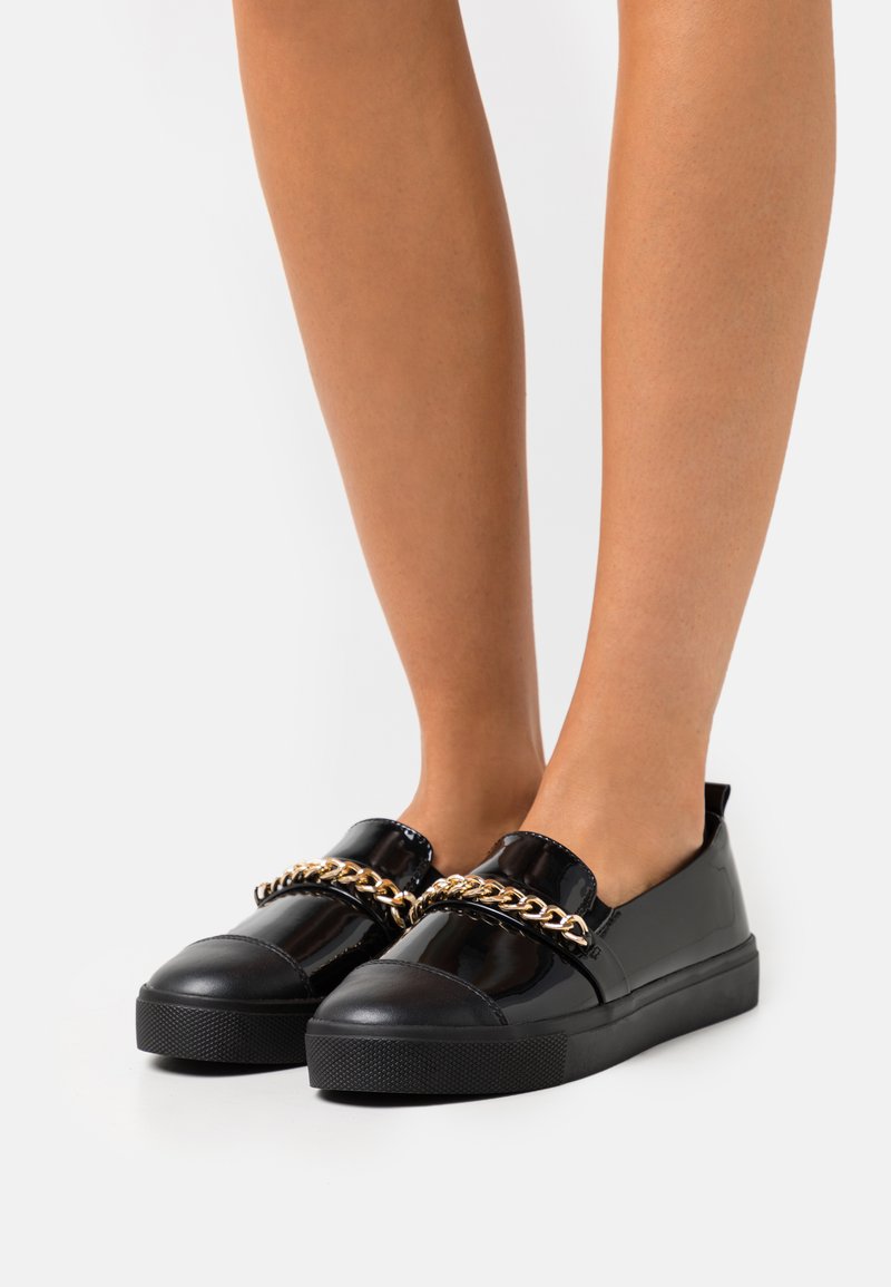 Black patent slip-on shoes with a thick black sole, featuring a gold chain detail across the front and a smooth, shiny texture.