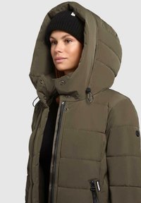 Vrouw in een olijfgroene gewatteerde winterjas met een hoog gewatteerd capuchon en een zwarte gebreide beanie voor een effen grijze achtergrond.