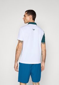 Camisa polo masculina, branca com colarinho azul-turquesa, de manga curta, e um logótipo bordado nas costas. Combinada com calções azuis, exibindo tatuagens.