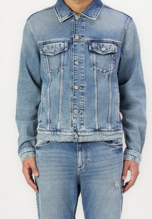 Mężczyzna ubrany w jasnoniebieską kurtkę jeansową z zapięciem na guziki i kieszeniami z klapami na piersiach, w zestawie z dopasowanymi jasnoniebieskimi jeansami.