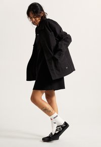 Veste noire surdimensionnée avec de gros boutons, robe noire en dessous, chaussettes Vans blanches et chaussures noires avec des accents métalliques et des semelles en caoutchouc.