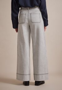 Grijze denim broek met wijde pijpen en een beschadigde zoom, twee achterzakken en zichtbare stiksels. Gepaard met zwarte enkellaarzen.