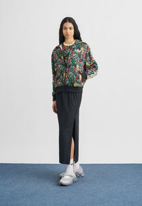 Schwarz gemusterte Jacke mit Reißverschluss und floralem Muster, Ärmeln und Taschen, kombiniert mit einem schwarzen Maxirock mit seitlichem Schlitz, dazu graue Sportschuhe.