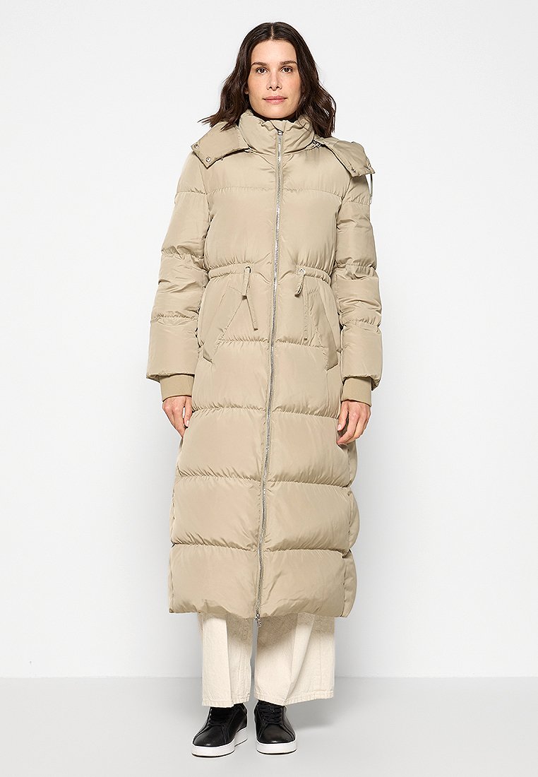 Manteau rembourré long beige avec une fermeture éclair sur le devant, des liens de taille réglables et un col haut. Présente des coutures en panneaux et est associé à des chaussures noires.