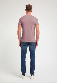 Kurzärmliges mauve T-Shirt mit Rundhalsausschnitt, kombiniert mit eng anliegenden blauen Jeans und weißen Sneakers, von hinten betrachtet.