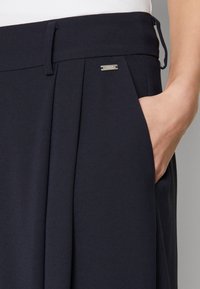 Laivasto sininen plissertu hame, jossa on sileä pinta, sivutaskut ja pieni metallikoriste vyötärökaistaleessa. Klassinen muotoilu, joka sopii erilaisiin tilaisuuksiin.