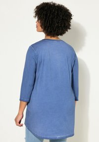 Túnica de denim azul con mangas tres cuartos, dobladillo ligeramente curvado y un corte holgado. Textura suave con costuras visibles.