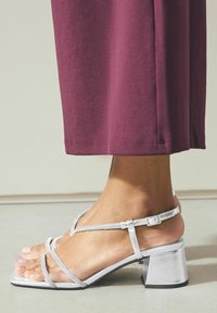 Silberne Sandalen mit dünnen Riemen und dekorativen Details, die eine quadratische Zehenpartie und einen strukturierten Blockabsatz zeigen. Kombiniert mit einem burgunderfarbenen Kleidungsstück.