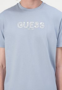 Vaaleansininen lyhythihainen t-paita, joka on valmistettu pehmeästä puuvillasta. Paidassa on valkoisella tekstillä "GUESS" ja sen alla selkeällä ja yksinkertaisella fontilla "EST. 1981".