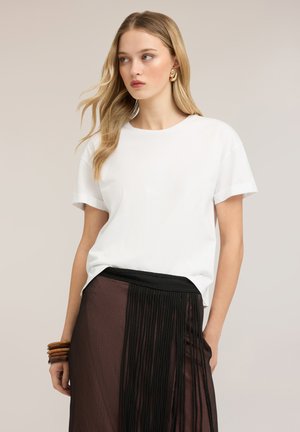 Ung kvinde med blondt hår iført en hvid t-shirt, plisseret sort og brun nederdel, guldbøjler og træarmbånd, der ser til siden.
