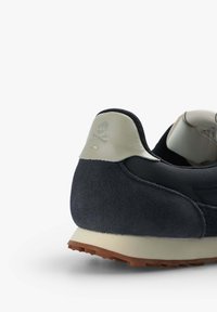 Mustat mokkanahka sneakerit, joissa on teksturoitu kantapää, joka esittelee pääkallon ja ristiluiden logon. Kumipohja on vaaleanruskea. Sileä kankainen sisustus.