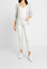 Ljusgrå stickad cardigan över en vit topp med spetskant, i kombination med ljusa raka jeans och vita sneakers.