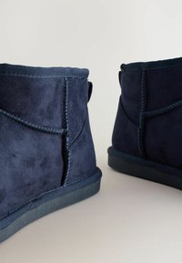 Navy plave suede čizmice s teksturiranim završetkom, s kontrastnim šavovima i ravnim gumiranim potplatom, prikazane sa stražnje strane.