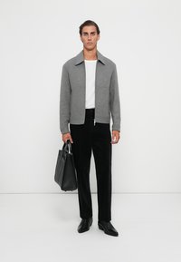 Pilka zip-up striukė, balta marškinėliai, juodos kelnės, odinė rankinė ir juodi batai; modelis stovi prieš vientisą baltą fono.