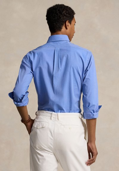 Bărbat văzut din spate, purtând o cămașă bleu cu mâneci lungi rulate și pantaloni albi, pe un fundal bej simplu.