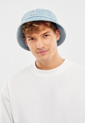 Mladý muž s kučeravými vlasmi, ktorý má na hlave svetlomodrú klobúk z rifľoviny a jednoducho bielu tričko s okrúhlym výstrihom, mierne sa usmievajúci na bielom pozadí.