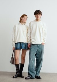 Νεαρή γυναίκα και άνδρας φορούντα αντίστοιχες κρεμ φούτερ, η γυναίκα φοράει τζιν φούστα και μπότες, ενώ ο άνδρας άνετο τζιν και λόφερ, στέκονται σε εσωτερικό χώρο.