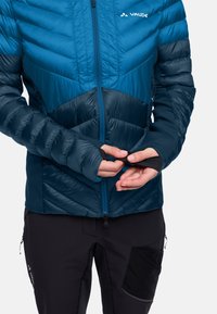 Una persona che regola il foro per il pollice della manica di una giacca trapuntata Vaude blu e navy, indossando pantaloni neri da esterno.
