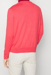 Korallfärgad stickad tröja med ribbad rund halsringning och långa ärmar, med en avsmalnande fåll. Bärs med ljusfärgade byxor.