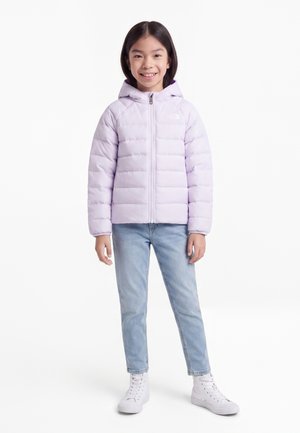 Pige smilende iført en lys lilla hættejakke, lyseblå jeans og hvide høje sneakers, stående mod en hvid baggrund.