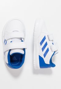 Vita atletiska skor med blå detaljer, med en syntetisk ovandel, två velcroremmar och texturerade sulor med en böjd design.