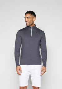 Szürke, hosszú ujjú, zipzáros póló, állógallérral és világoskék cipzárral; Calvin Klein logóval az azonos színű cérnával. Alatta fehér rövidnadrág.