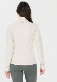 Helles beige, langärmliges Sportoberteil mit hohem Kragen, weichem Material und flachen Nähten. Mit einem Logodetail am hinteren Kragen.